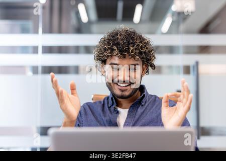 Photo en gros plan d'un jeune Indien assis à son bureau dans le bureau, souriant et regardant l'écran de son ordinateur portable joyeusement. Banque D'Images
