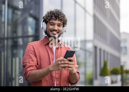 Portrait d'un jeune Indien portant des écouteurs blancs tenant un téléphone et souriant à la caméra. Banque D'Images