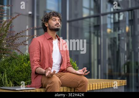 Un jeune Indien calme et détendu est assis près d'un bâtiment sur un banc dans une position de lotus avec les yeux fermés et médite. Banque D'Images