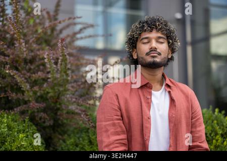 Photo en gros plan d'un jeune Indien en chemise rouge assis sur un banc dehors, les yeux fermés et reposant paisiblement. Banque D'Images