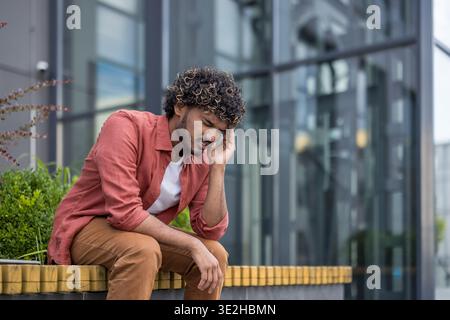 Jeune homme indien fatigué assis sur un banc près d'un immeuble de bureaux et tenant sa tête avec sa main. Banque D'Images