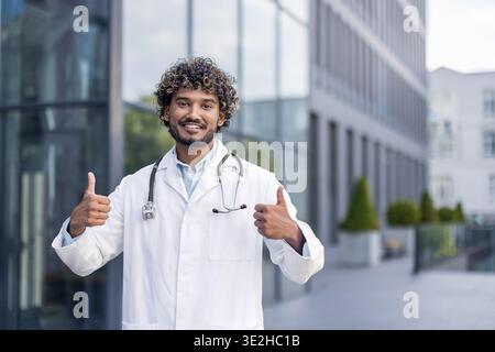Portrait d'un jeune médecin indien souriant, étudiant debout à l'extérieur de la clinique dans un manteau blanc et montrant le super signe avec ses doigts à la caméra. Banque D'Images