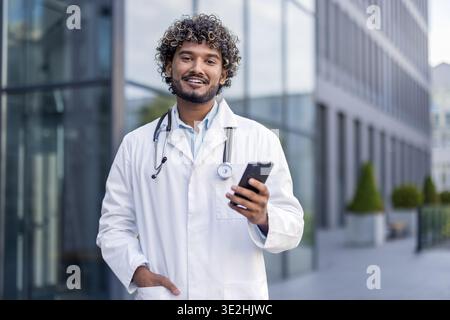 Portrait d'un jeune médecin indien souriant debout dehors, tenant un téléphone dans sa main et regardant la caméra. Banque D'Images