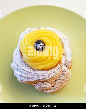Zeppola di San Giuseppe représentant la pâtisserie italienne traditionnelle, remplie de crème anglaise riche et garnie d'une cerise noire brillante, élégamment saupoudrée de sucre en poudre sur une assiette vert clair Banque D'Images