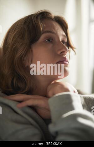 Verticale de jeune femme adulte caucasienne assise à l'intérieur reposant le menton sur les mains regardant soigneusement dans la distance montrant l'expression faciale contemplative soulignant l'émotion et l'introspection Banque D'Images