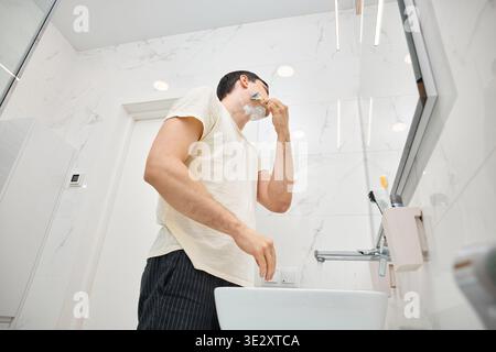 Homme rasant le visage pendant la routine d'hygiène du matin dans la salle de bain moderne blanche Banque D'Images
