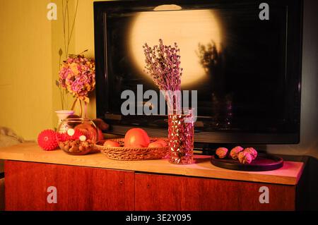Une collection d'articles décoratifs comprenant un arrangement floral, lavande, fruits dans un panier, noix, et un écran de télévision avec un reflet Banque D'Images