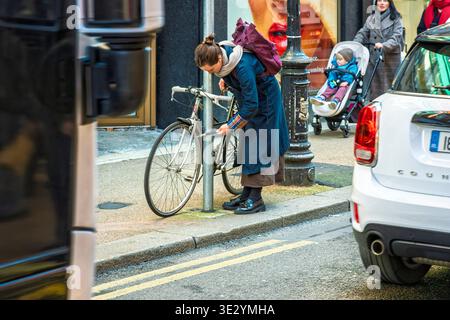 Femme verrouillant un vélo à un poteau dans une rue animée du centre-ville de Dublin avec piétons et circulation. Banque D'Images