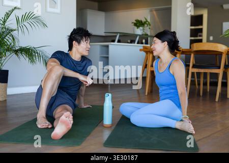 Couple diversifié assis et bavardant dans des vêtements athlétiques sur des tapis verts à la maison avec bouteille bleue Banque D'Images
