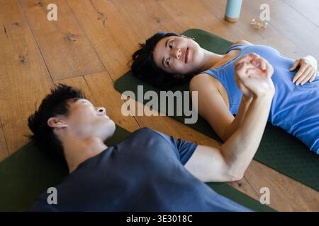 Couple asiatique couché, tenant les mains sur des tapis verts à la maison dans des vêtements de sport, bouteille d'eau visible Banque D'Images