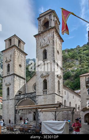 Kotor, Monténégro - 20 mars 2026 : vue en bas angle des tours romanes de la cathédrale Saint Tryphon et le drapeau national du Monténégro agitant dans le Banque D'Images