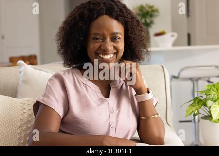 Femme africaine adulte assise sur un canapé beige clair à la maison souriant, portant un chemisier rose et smartwatch Banque D'Images