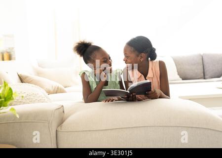 Filles africaines enfant-adolescentes se prélassant sur un canapé dans le salon tenant livre ouvert, lecture et liaison Banque D'Images