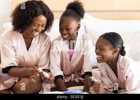 Mère de famille afro-américaine avec adolescent, filles d'enfant sur le lit à l'aide de vernis à ongles dans des robes roses Banque D'Images