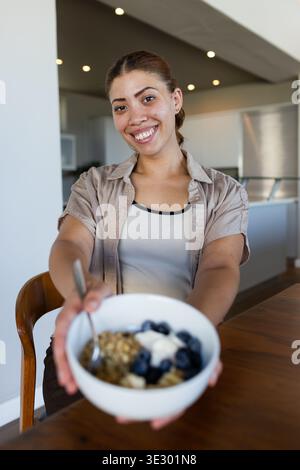 Femme afro-américaine souriante et tenant un bol blanc de myrtilles granola et de yaourt dans la cuisine Banque D'Images