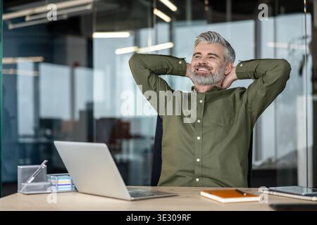 Heureux homme d'affaires mature penché en arrière dans sa chaise, souriant avec les mains derrière la tête, prenant une pause bien méritée à son bureau dans un bureau contemporain ouvert Banque D'Images