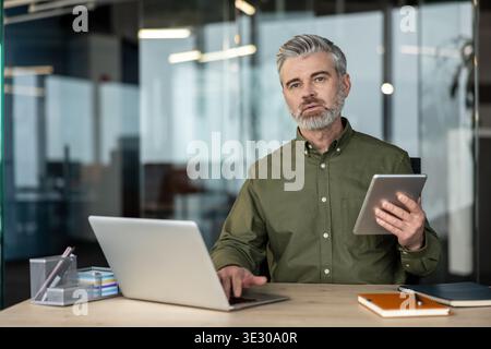 Homme d'affaires mûr avec les cheveux gris et la barbe est assis à un bureau moderne, en utilisant un ordinateur portable et une tablette pour le travail à distance et la communication en ligne, axé sur la planification et la stratégie Banque D'Images