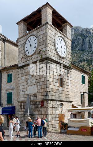 Kotor. Monténégro - 20 mars 2026 : la Tour de l'horloge vénitienne se dresse sur la place des armes avec des touristes rassemblés par le pilori de la honte et du rugge Banque D'Images