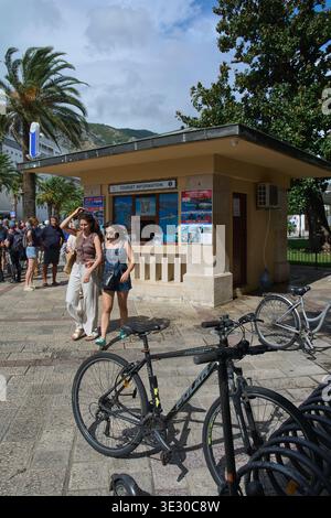Kotor. Monténégro - 20 mars 2026 : deux jeunes femmes passent devant un kiosque d'information touristique au port de Kotor près de vélos stationnés et de palmiers Banque D'Images