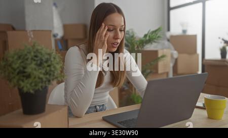 Femme avec la main sur la joue regardant l'ordinateur portable dans le bâtiment entouré de boîtes en mouvement emballées, plante, et tasse sur la table ; stress de jour en mouvement. Banque D'Images