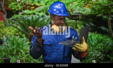 Homme ouvrier de la construction dans un casque bleu tenant un ordinateur portable et une clé, haussant les épaules avec la main gantée dans la forêt ; dépannage de frustration. Banque D'Images