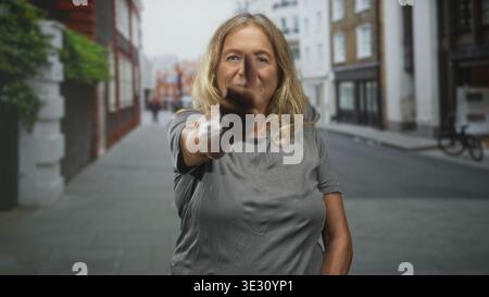 Femme âgée aux cheveux blonds pointe doigt directement à la caméra sur une rue urbaine bordée de bâtiments et de vélos ; confiance. Banque D'Images