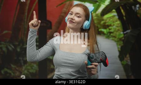 Jeune femme rousse tenant une manette de jeu sans fil et portant un casque d'oreille de chat bleu pointe l'index vers le haut dans la forêt ; ludique. Banque D'Images