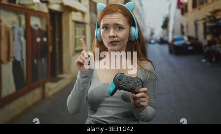 Femme rousse tenant une manette de jeu et pointant l'index tout en portant des écouteurs bleus dans une rue ; focalisation contemplation. Banque D'Images