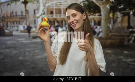 Femme souriant et tenant un canard en caoutchouc jaune tout en donnant les pouces sur une rue historique avec la façade du bâtiment ; jeu joyeux. Banque D'Images
