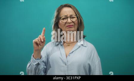 Femme senior pointe le doigt avec index relevé et main visible tout en portant des lunettes et une chemise bleu clair dans le studio ; avertissement de désapprobation. Banque D'Images
