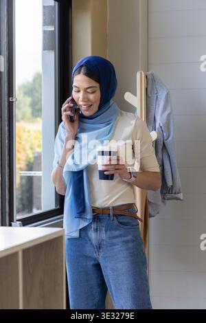 Femme en hijab bleu debout par la fenêtre à la maison tenant le café et smartphone, smartwatch visible Banque D'Images