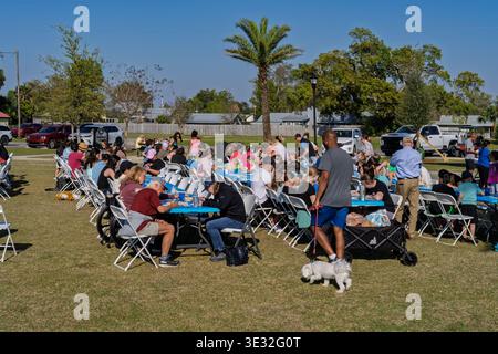 Lynn Haven, Floride, États-Unis. 20 mars 2026. Des dizaines de membres de la communauté se rassemblent autour de longues tables pliantes bleues à Sheffield Park pour peindre sur des toiles fournies gratuitement lors de l’événement Art in the Park parrainé par la ville. Les familles, les amis et les résidents locaux de tous âges et de tous niveaux de compétence profitent d’un après-midi ensoleillé exprimant leur créativité ensemble dans un magnifique cadre extérieur. Au premier plan, un homme en chemise grise promène un petit chien blanc devant la scène animée tandis que les participants se concentrent sur la création de leur prochain grand chef-d'œuvre. Crédit : Tosh Persons / Alamy Live News Banque D'Images