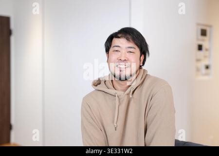 Sourire chaleureux d'un homme détendu et décontracté. Un éclairage tamisé met en valeur l'expression douce et le sweat à capuche confortable. Le cadre accueillant suggère le confort et l'approa Banque D'Images