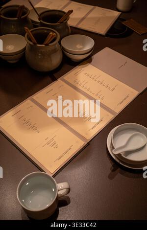 La table aux tons chauds contient des bols et des tasses en céramique. Les menus manuscrits reposent à plat sur une surface en bois. Les pots en céramique robustes contiennent des bâtons en bois fins. Simple Banque D'Images