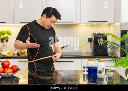 L'homme asiatique en surpoids dans la cuisine à la maison mesure la taille avec du ruban adhésif et semble frustré par le gros ventre après le gain de poids. Les capsules de régime sur la table suggèrent la recherche pour le changement de routine de santé de soutien amincissant. Banque D'Images