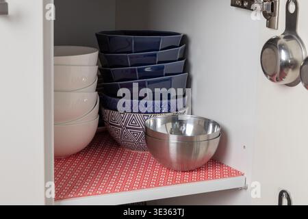 Vue de face d'une armoire de cuisine rangée doublée d'un tapis rouge, contenant des bols en céramique empilés et un bol en métal. La disposition propre met en évidence l'ordre et la mijoté Banque D'Images