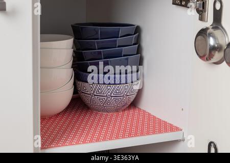 Vue de face d'une armoire de cuisine rangée doublée d'un tapis rouge, contenant des bols en céramique empilés. L'arrangement propre met en évidence l'ordre et l'organisat de la maison simple Banque D'Images