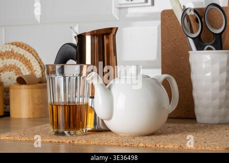 Théière en céramique blanche et verre transparent rempli de thé placé sur un tapis de liège à côté d'une machine à expresso en cuivre. Intérieur de cuisine chaleureux avec des textures en bois c Banque D'Images