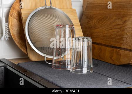 Verres à boire transparents placés à l'envers sur un tapis de séchage près de planches à découper en bois et d'un tamis métallique. La scène lumineuse de la cuisine met en évidence la propreté, o Banque D'Images