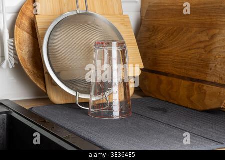 Verres à boire transparents placés à l'envers sur un tapis de séchage près de planches à découper en bois et d'un tamis métallique. La scène lumineuse de la cuisine met en évidence la propreté, o Banque D'Images
