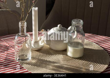 Ce cliché en grand angle capture un petit-déjeuner confortable et paisible. Sur un napperon tissé rond, une théière en céramique blanche se trouve à côté d'un bo en verre Banque D'Images