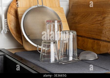 Verres à boire transparents placés à l'envers sur un tapis de séchage près de planches à découper en bois et d'un tamis métallique. La scène lumineuse de la cuisine met en évidence la propreté, o Banque D'Images