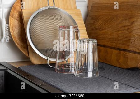 Verres à boire transparents placés à l'envers sur un tapis de séchage près de planches à découper en bois et d'un tamis métallique. La scène lumineuse de la cuisine met en évidence la propreté, o Banque D'Images