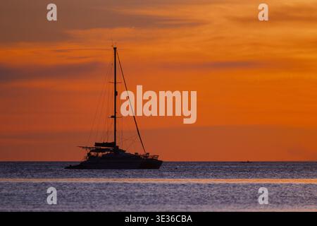 Une silhouette de voilier repose sur les eaux calmes de l'océan sous un coucher de soleil orange vif, évoquant la tranquillité, la solitude et l'ambiance tropicale de soirée idéale pour trav Banque D'Images