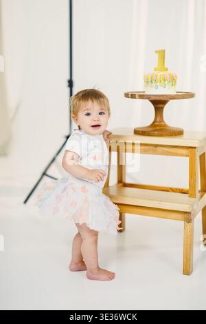 Un enfant se tient près d'une petite table avec un gâteau d'anniversaire mettant en vedette le numéro un. La scène se déroule dans un studio avec un éclairage doux, en se concentrant sur le c Banque D'Images