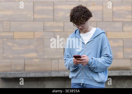 Adolescent mâle debout par le mur de pierre tan, tenant le téléphone rouge, portant des lunettes et des jeans, espace de copie Banque D'Images