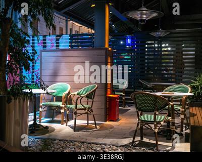 Le patio extérieur moderne d'un restaurant ou d'un bar est doté de chaises tressées vertes, de petites tables et de cloisons en bois éclairées par des lampes chaudes A. Banque D'Images