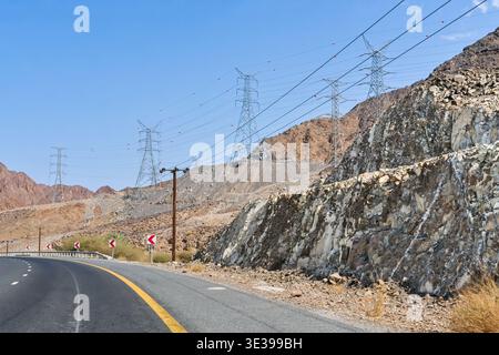Tours de transmission d'énergie à haute tension et lignes électriques longeant la route de montagne dans le paysage désertique des Émirats arabes Unis. Banque D'Images