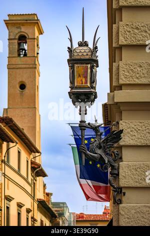 Support de lanterne dragon en fer orné sur un bâtiment historique en pierre sur une rue, clocher et drapeaux européens en arrière-plan à Florence, en Italie Banque D'Images