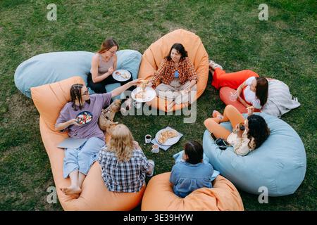 Groupe d'amis se relaxant sur des sacs de haricots colorés à l'extérieur, partageant pizza et boissons dans une arrière-cour décontractée se réunissant sous la douce lumière de l'été. Vue de dessus Banque D'Images